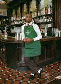 A model leaning against a bar wearing the Guinness x Percival forest green apron over a white shirt and pinstripe trousers.
