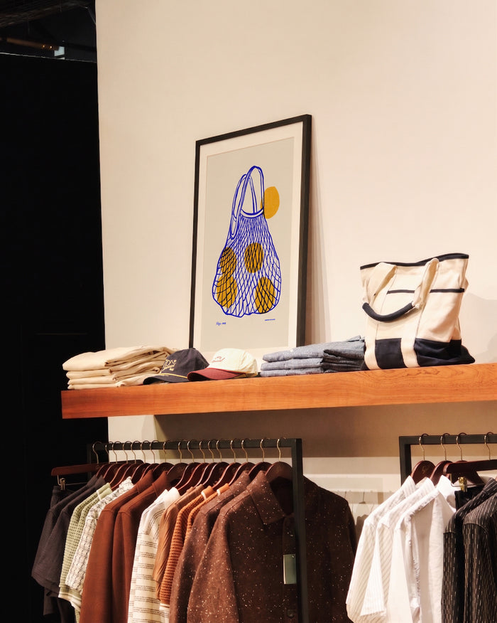 The framed Arancia Print from Percival displayed on a wooden shelf above clothing rails in a store.