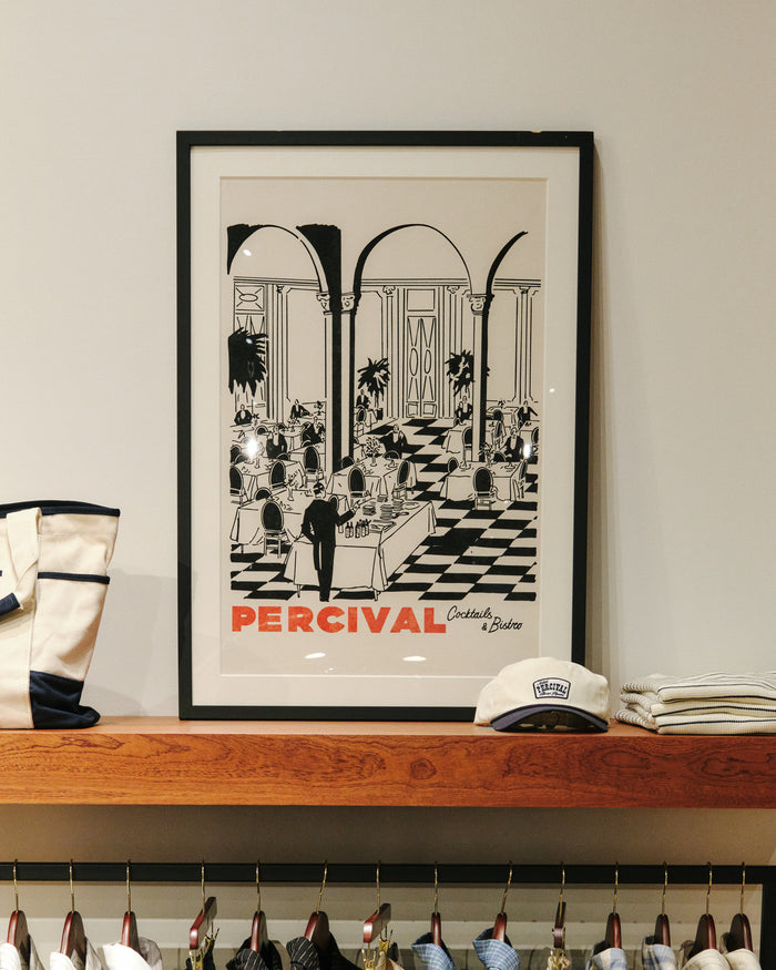 The Percival Bistro print in a black frame on a wooden shelf next to a tote bag and cap.