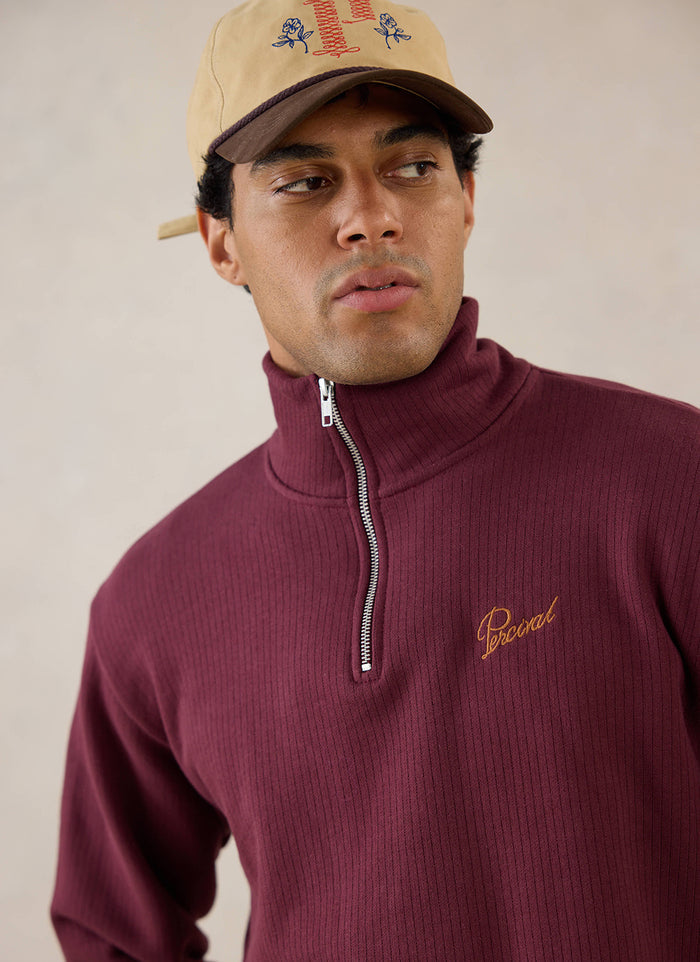 A close-up of a model wearing the burgundy Percival Conrad Funnel Neck 1/4 Zip Jumper and a cap, showing the collar detail.