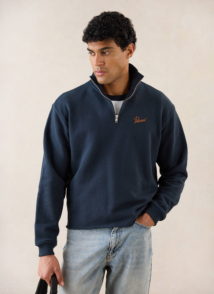 A model wears the navy Percival Conrad 1/4 zip cotton crew neck with orange embroidery, styled with blue jeans in a studio.