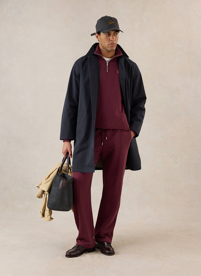 A model wears the merlot Percival Conrad trackpants, styled with matching jumper, dark coat, cap, and holdall.