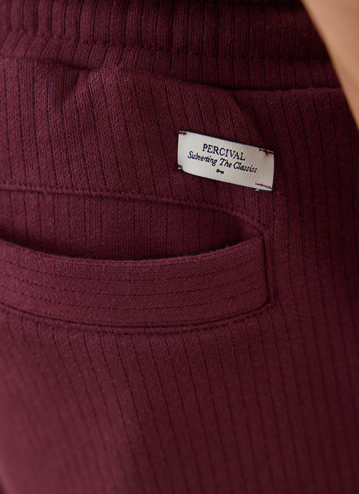 A close-up of the burgundy cotton trackpants from Percival, showing the branded label on the back pocket.