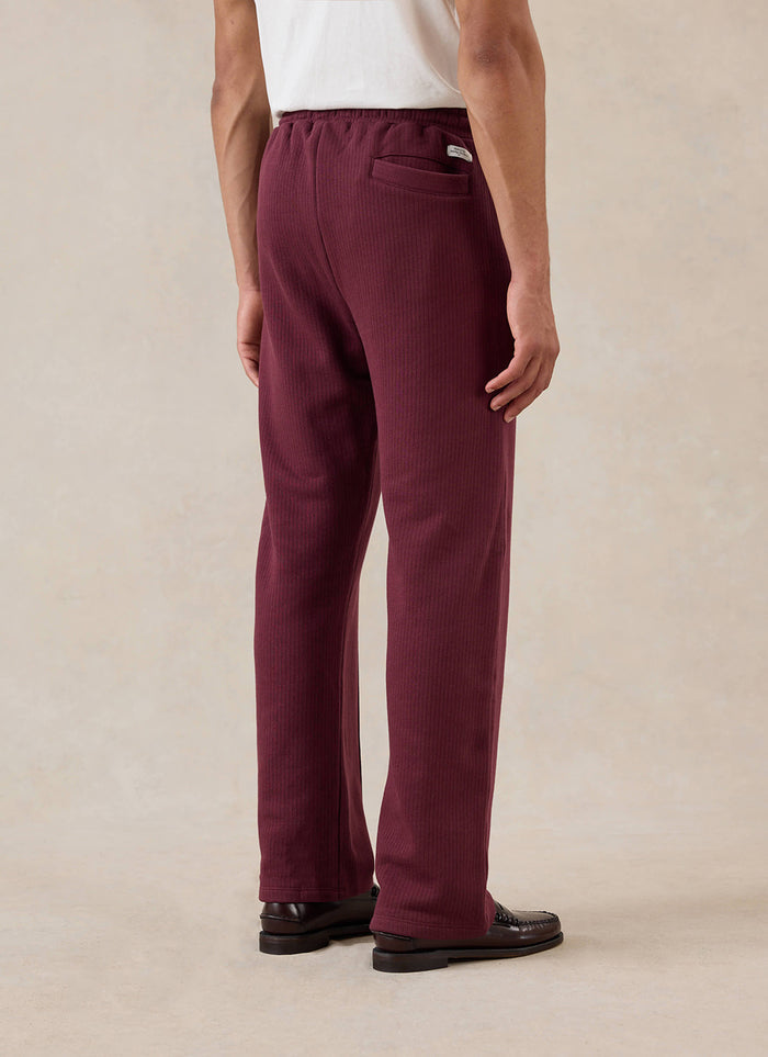 A model wears the merlot Percival Conrad straight leg trackpants from behind, paired with a white t-shirt and loafers.