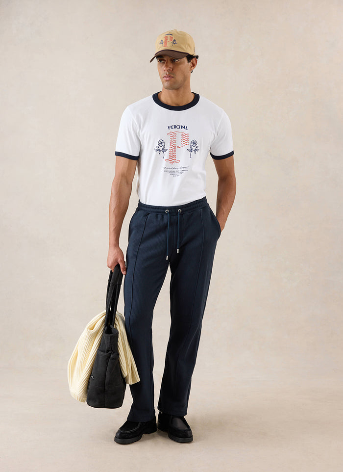 A model wears navy Percival Conrad trackpants, styled with a white ringer T-shirt, cap, tote bag and black shoes.