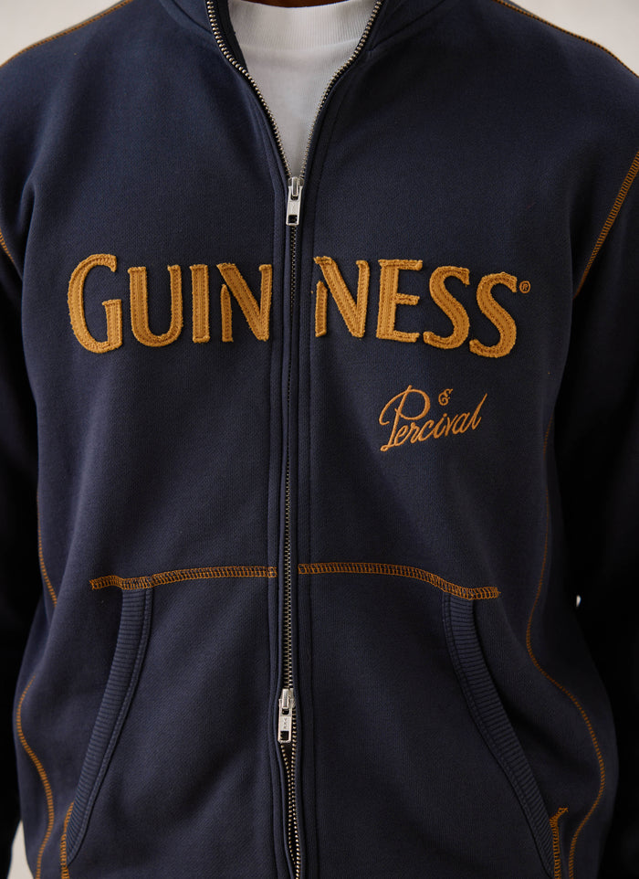 A close-up of the navy blue Guinness x Percival zip-up sweatshirt, showing the gold appliqué branding and contrast stitching.