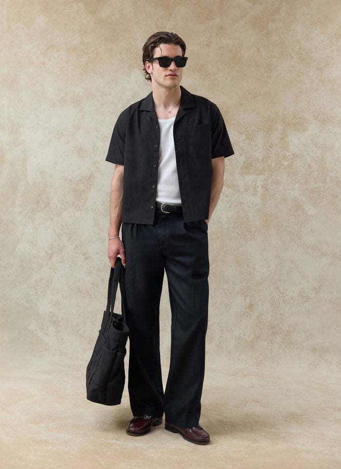 Percival black cotton Daniel Boxy Cuban Shirt worn open with a white vest and dark trousers in a studio setting.