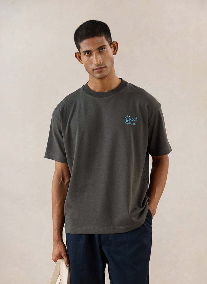A model wears the charcoal Percival Dawson boxy t-shirt, featuring light blue embroidery on the chest.
