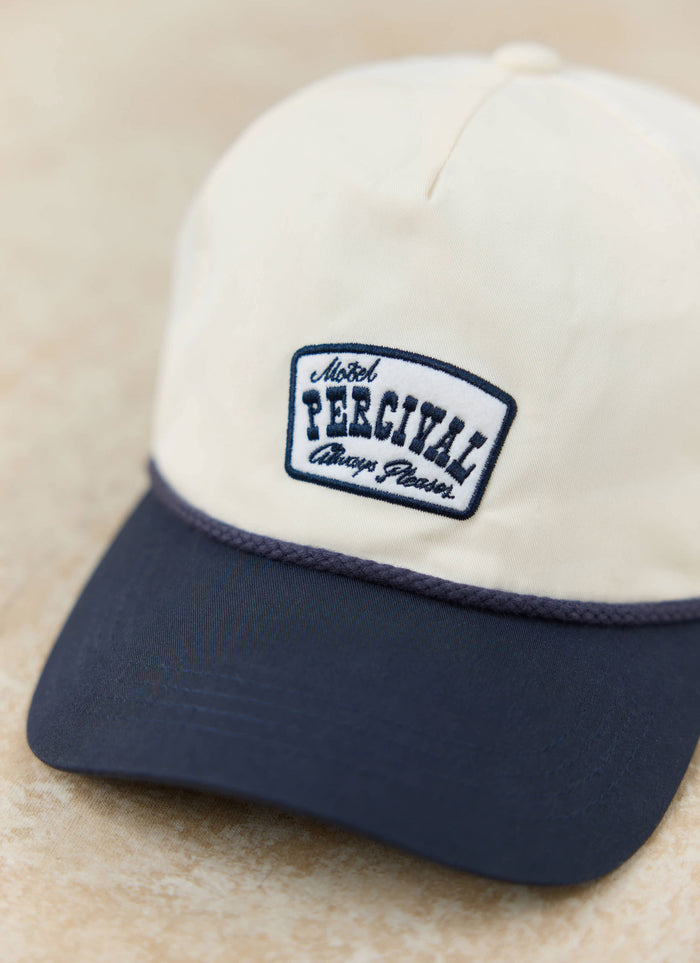 Close-up of the cream Percival Donovan soft trucker cap, featuring a navy brim and an embroidered motel-inspired patch.