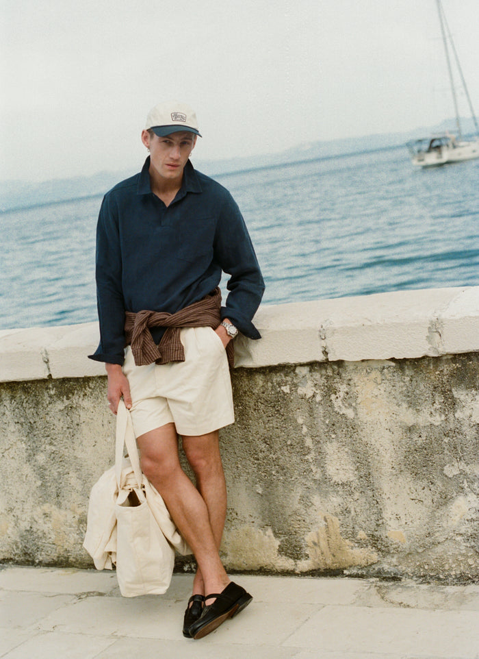 Model wears the Percival Donovan Soft Trucker Cap in cream cotton and navy with an embroidered patch in a coastal setting.