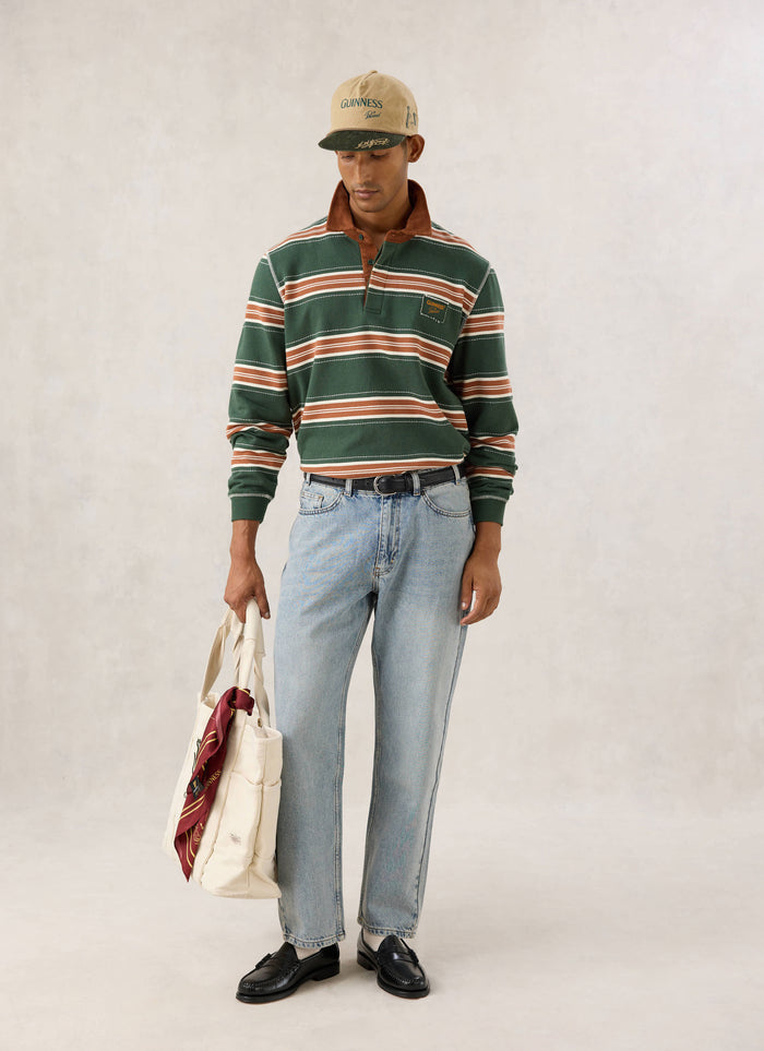 Model wearing the Guinness x Percival striped rugby polo shirt in forest green with a cord collar and cap.
