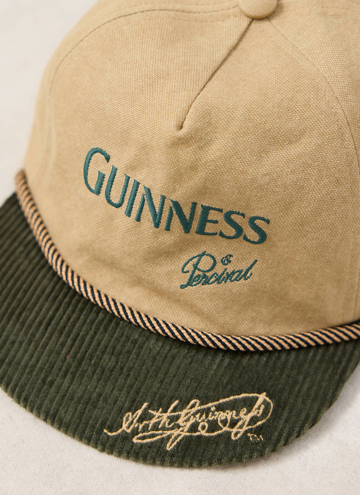 Close-up of the beige Guinness x Percival short peak cap, showing the green corduroy brim and front embroidery.