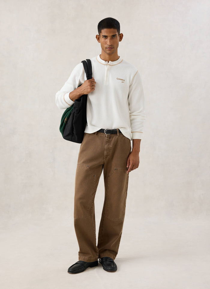 Model wearing the cream Guinness x Percival waffle henley long sleeve shirt with brown trousers and a black bag.