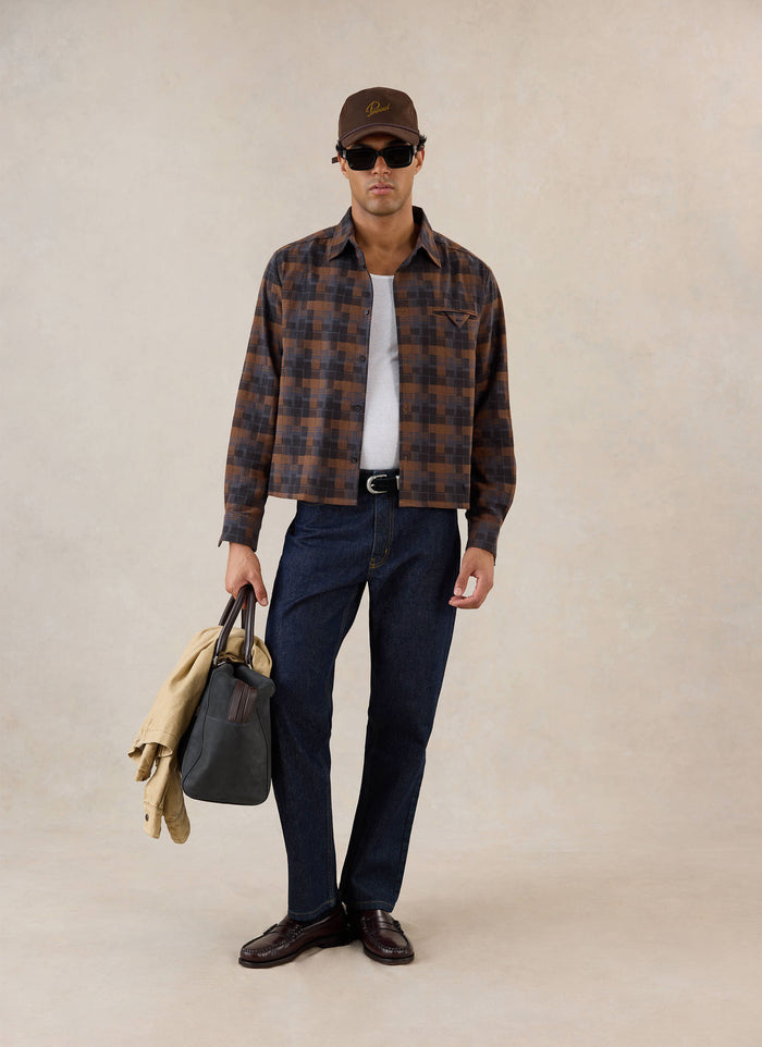 A model wears the brown check Percival shirt over a white vest, styled with dark jeans, sunglasses, cap, and a carry bag.