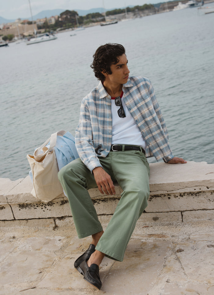 Percival Jesse cropped shirt in blue check linen-cotton, styled with sage trousers on a model sitting by the sea.