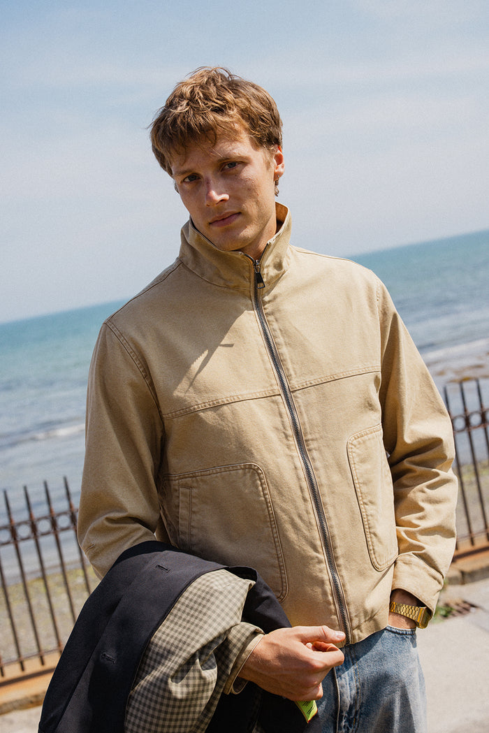 A model wears the beige cotton Percival Jackson Funnel Neck Harrington Jacket by the seaside.