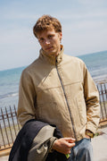 A model wears the beige cotton Percival Jackson Funnel Neck Harrington Jacket by the seaside.