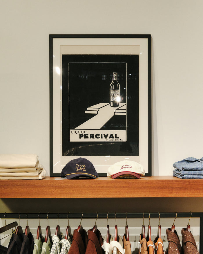 Framed Liquor print from Percival displayed on a wooden shelf above clothing rails in a store.