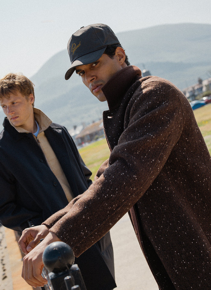 A model wearing the charcoal Percival Luther soft trucker cap and brown fleck Minnesota overcoat outdoors.