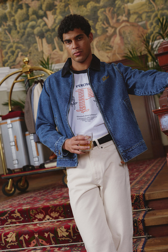 A model wearing a Percival mid wash blue denim zip jacket, white ringer t-shirt, and cream jeans indoors.