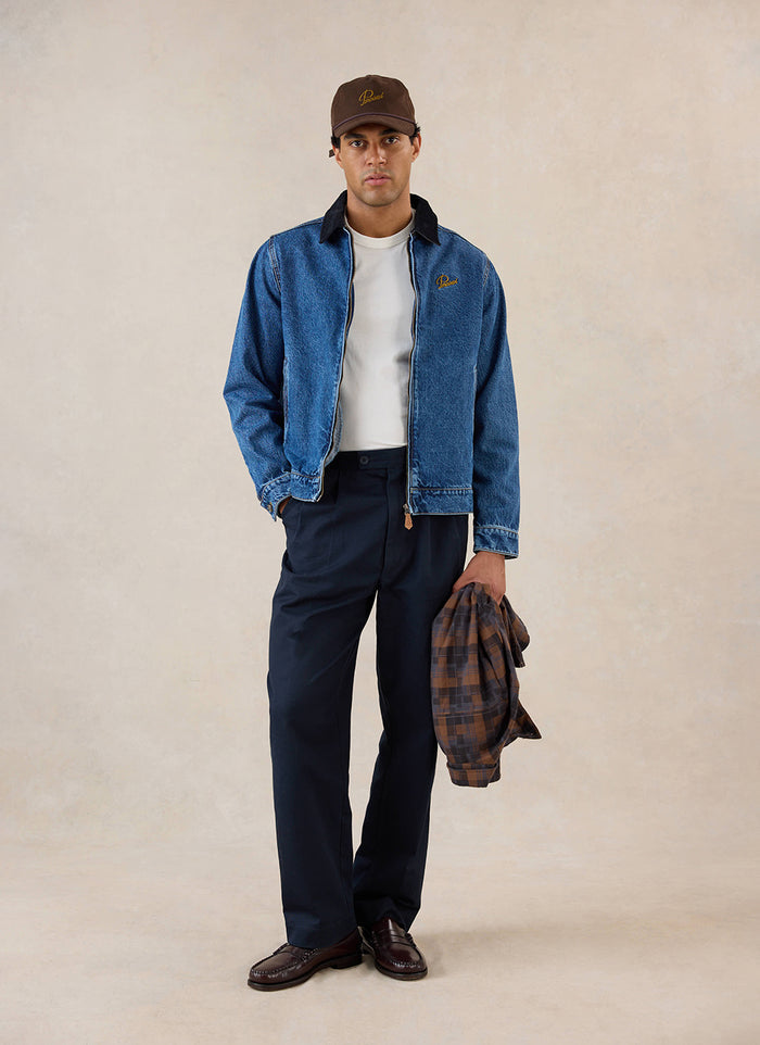 A model wears the mid wash blue Percival Mackey Zip Jacket, styled with a white tee, dark trousers and a brown cap.