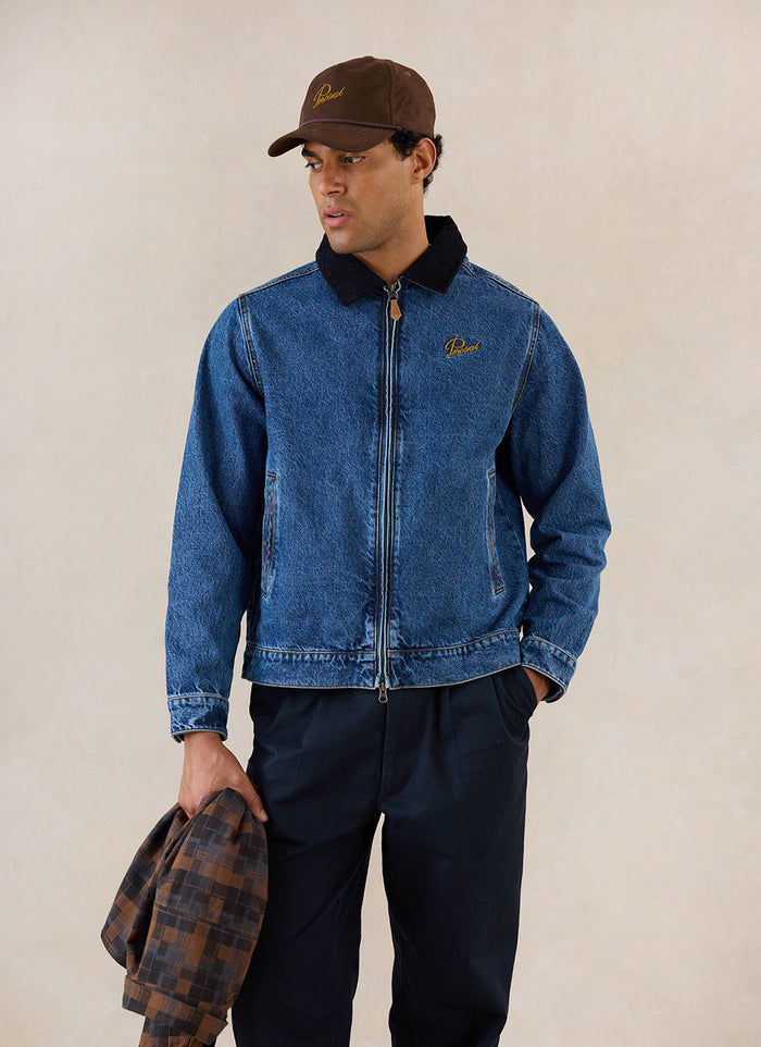 A model wears the mid wash blue Percival Mackey Zip Jacket in denim, styled with dark trousers and a brown cap.