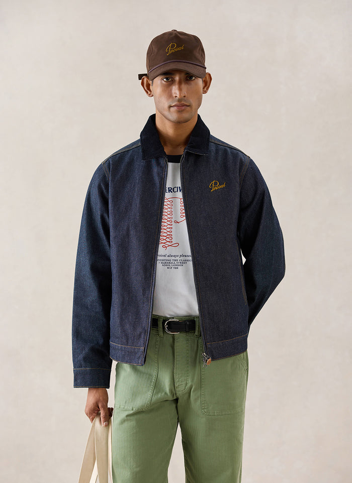 A model wears the raw indigo Percival Mackey Zip Jacket, styled with a graphic t-shirt and green trousers.