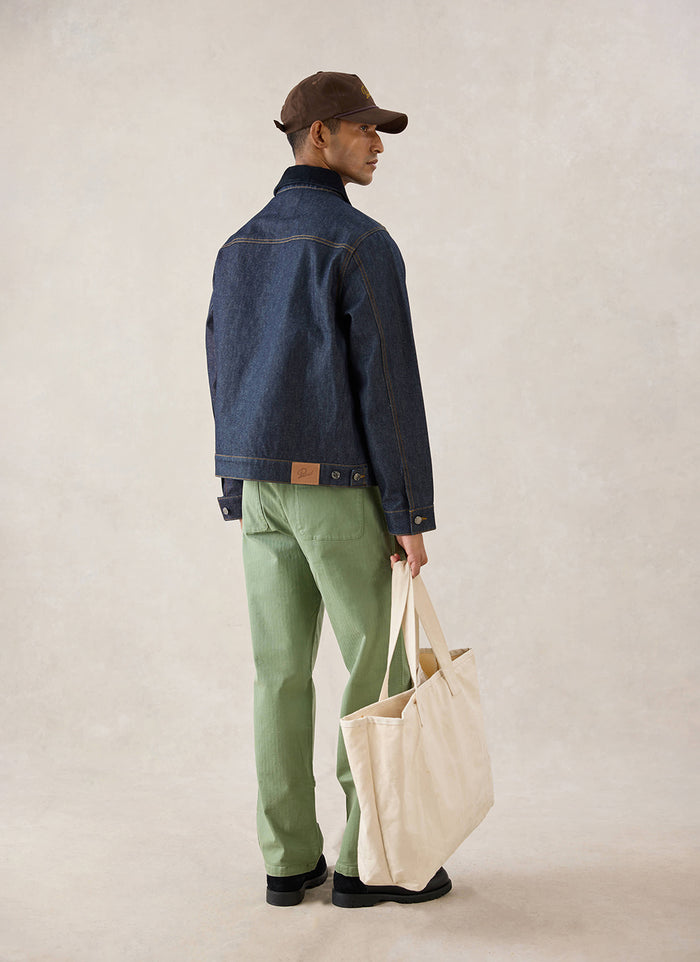 A model wears the raw denim Mackey Zip Jacket from Percival from behind, paired with green trousers and carrying a tote bag.