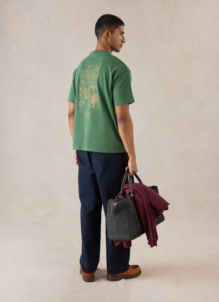 A model wears the forest green Percival Marley Boxy T-Shirt, showing the back graphic print while holding a dark holdall.