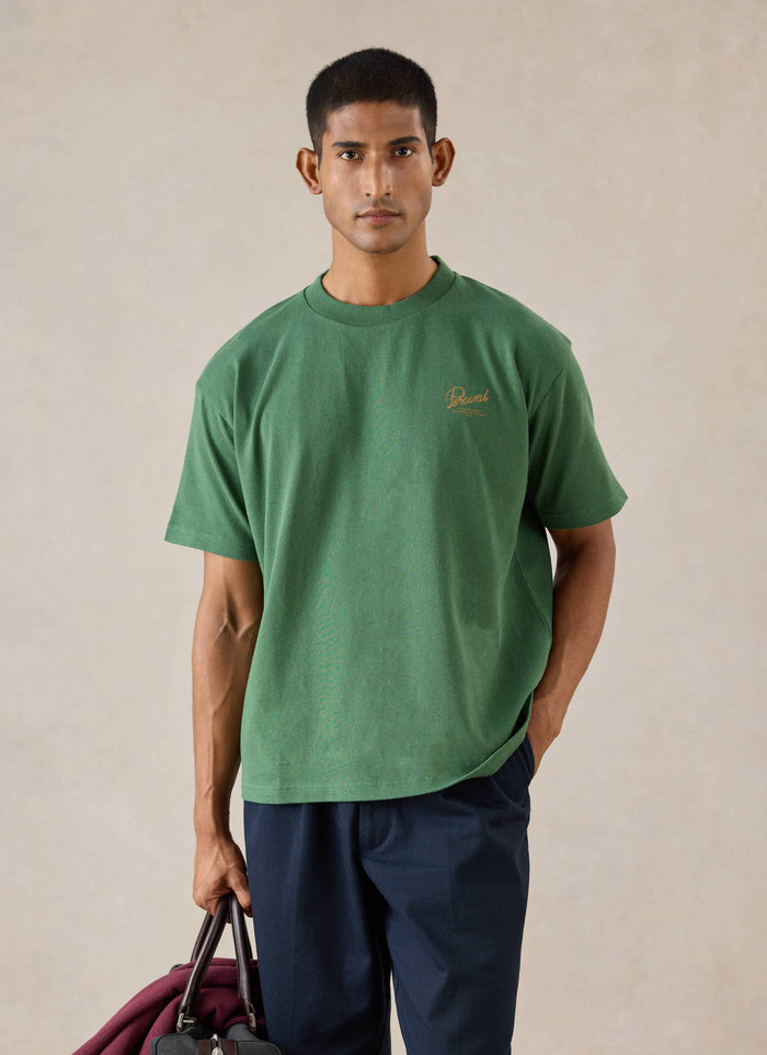 A model wears the forest green Percival Marley boxy t-shirt in organic cotton, styled with dark trousers.