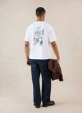 A model from behind wears the white Percival Marley boxy T-shirt featuring a dinner party back graphic and dark trousers.