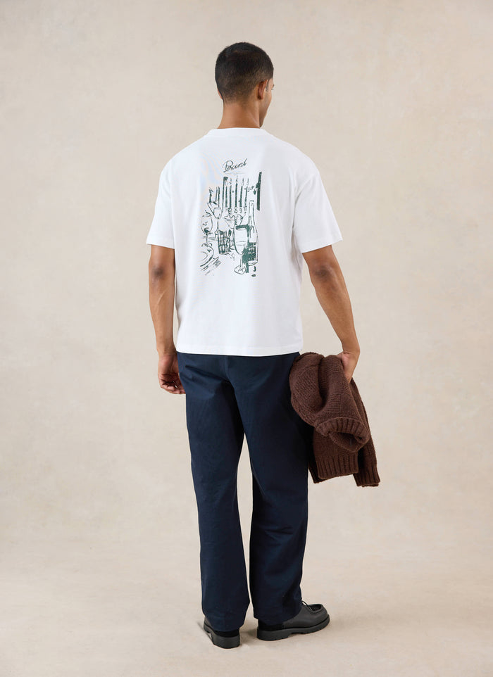 A model from behind wears the white Percival Marley boxy T-shirt featuring a dinner party back graphic and dark trousers.