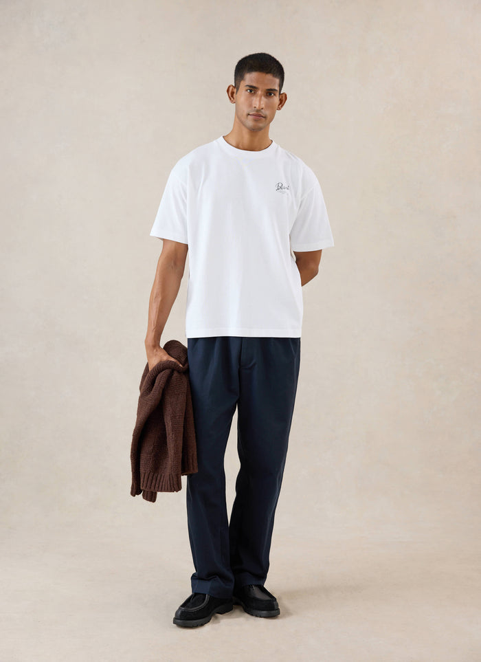 A model wears the white Percival Marley boxy t-shirt, styled with dark trousers and holding a brown knit jumper in a studio.