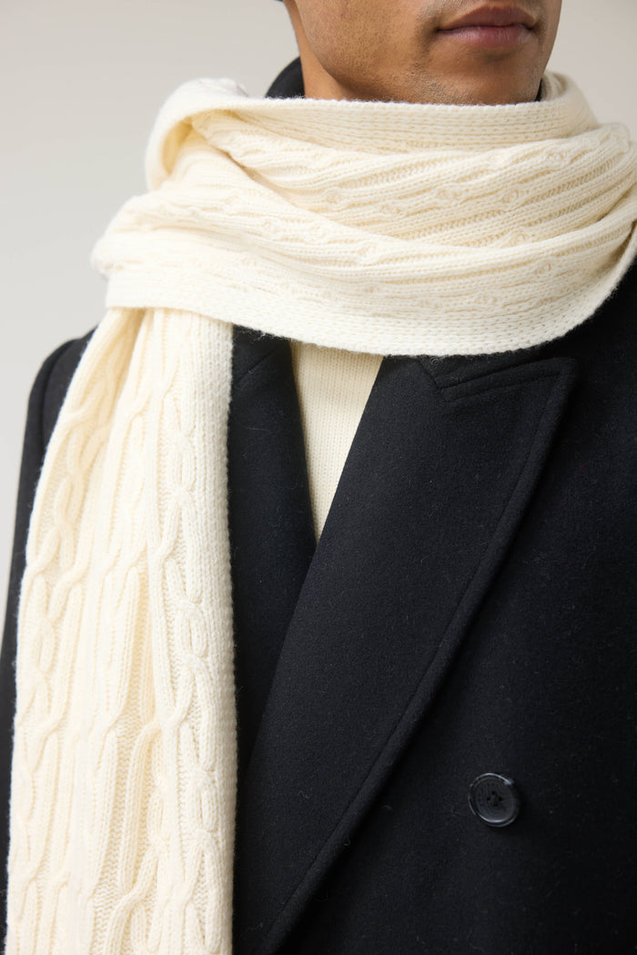 A close-up of a man wearing the ecru wool Melville Cable Knit Scarf from Percival over a dark coat.