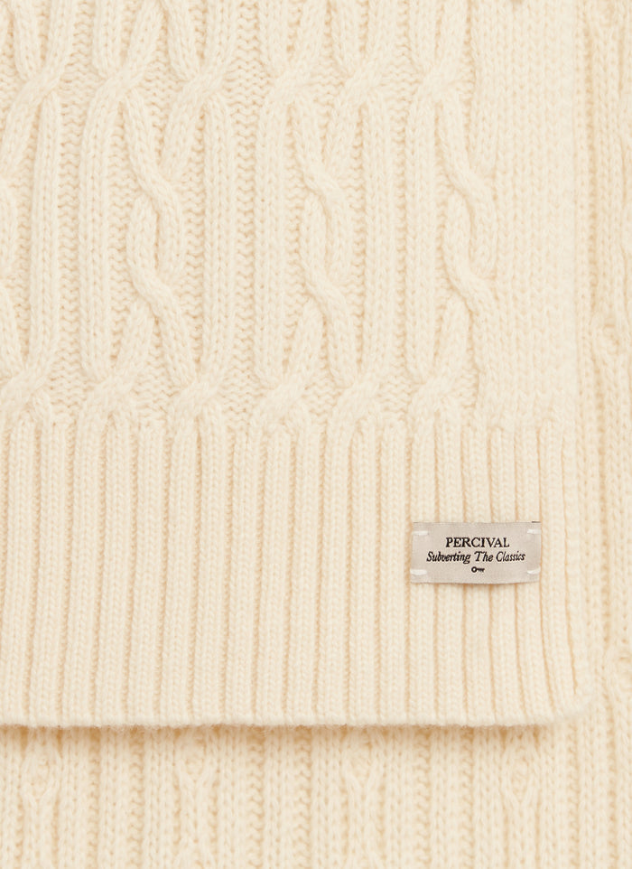 A close-up of the Percival brand label on the Melville Cable Knit Scarf in ecru, showing the detailed wool-blend cable knit.
