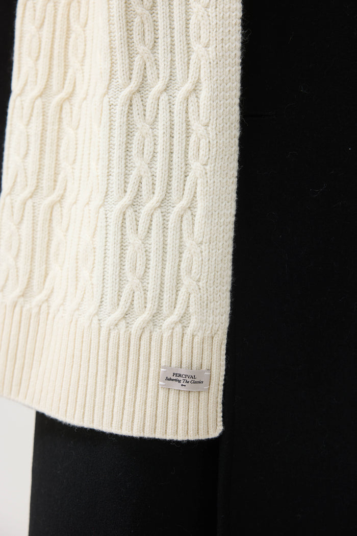 A close-up of the Percival Melville Cable Knit Scarf in ecru, showing the diagonal cable pattern and branded label.