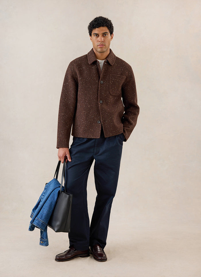 A model wears the brown fleck boxy overshirt from Percival with dark trousers and a black tote bag in a studio setting.