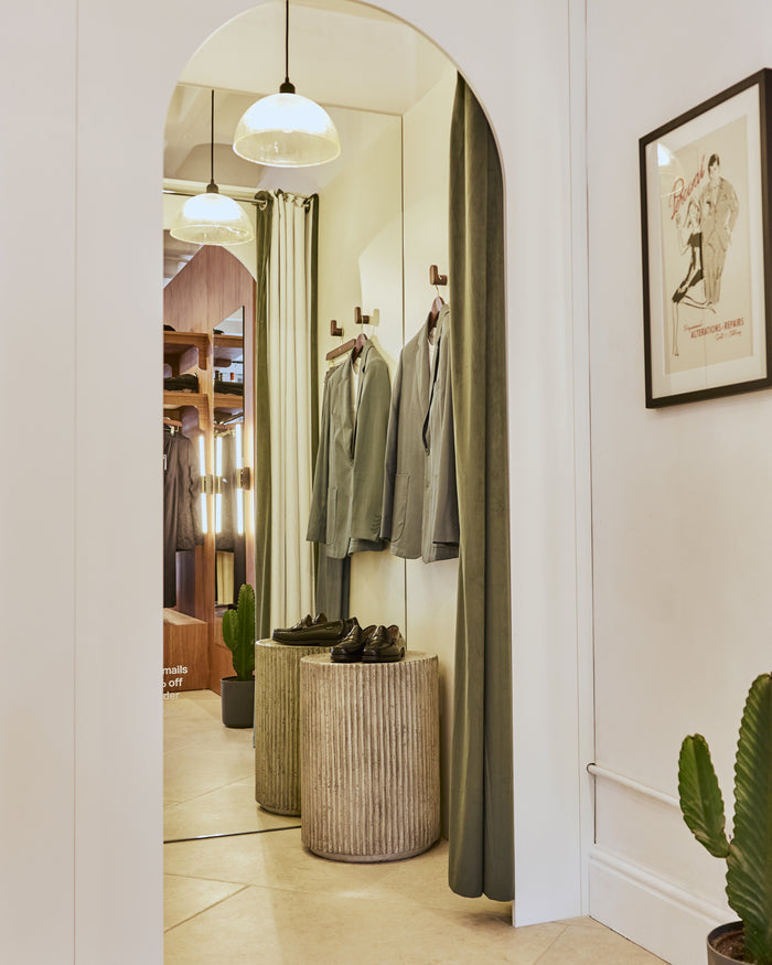 The Perci Alterations Print hanging on a wall in a Percival store interior, featuring hanging jackets and loafers.