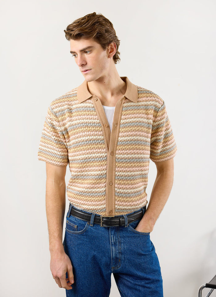 A male model wears the Percival Sari Stripe Knitted Shirt in ecru, styled open over a vest with blue jeans.