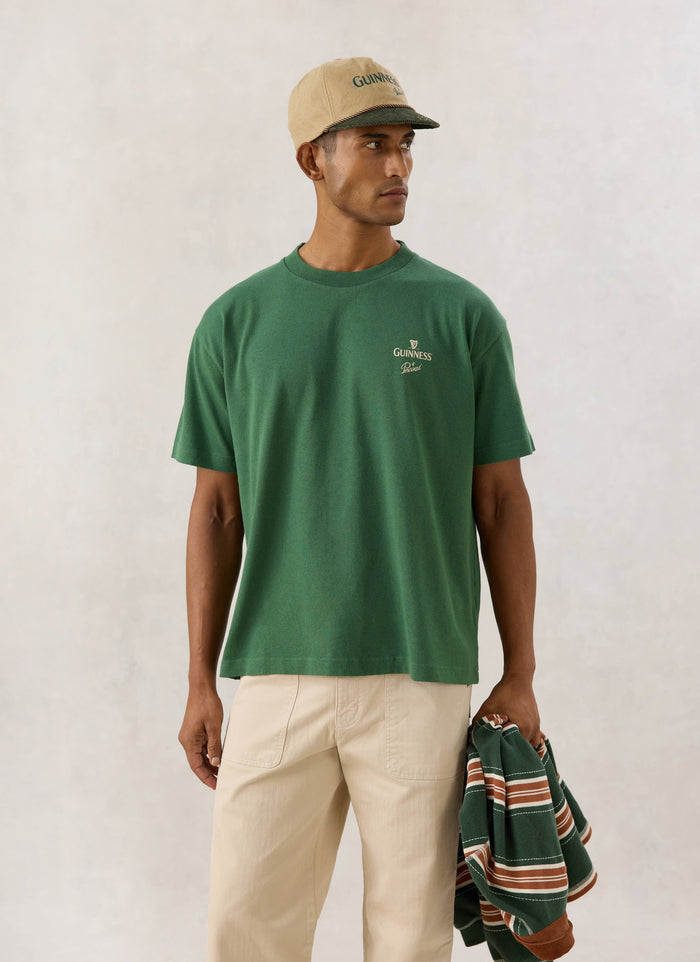 Model wearing the Guinness x Percival forest green organic cotton t-shirt with front chest branding and matching cap.