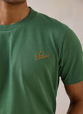 Close-up of a model wearing a forest green organic cotton Percival T-shirt with contrast script chest embroidery.