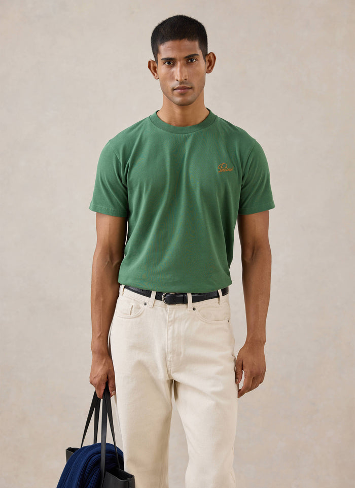 A model wears the forest green Percival Script Embroidered T-Shirt, styled with cream trousers and holding a tote bag.