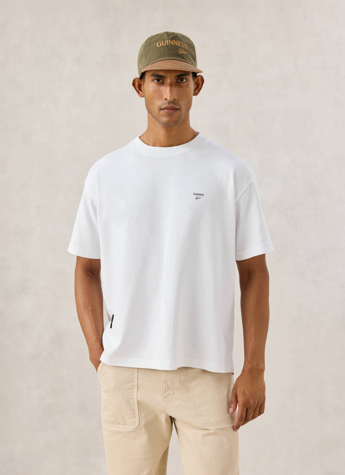 Front view of a model wearing the white Guinness x Percival Soloist oversized t-shirt and a branded cap.