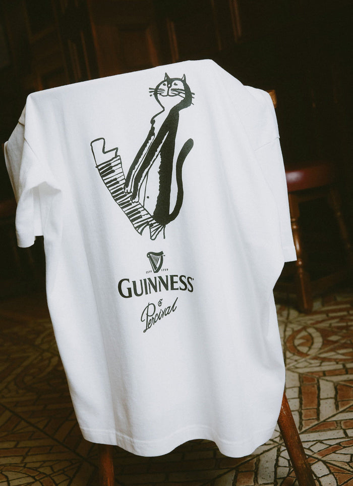 The Percival x Guinness Soloist T-shirt in white, draped over a wooden chair, showcasing the cat at the piano back print.