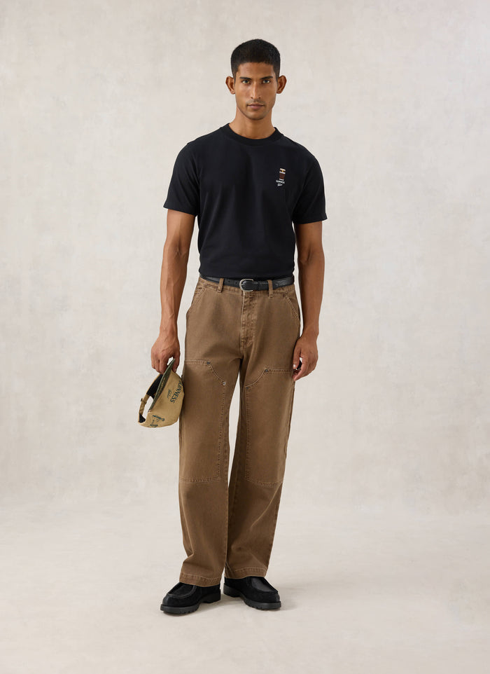 Model wearing the black Guinness x Percival stout t-shirt with chest embroidery, paired with brown trousers.
