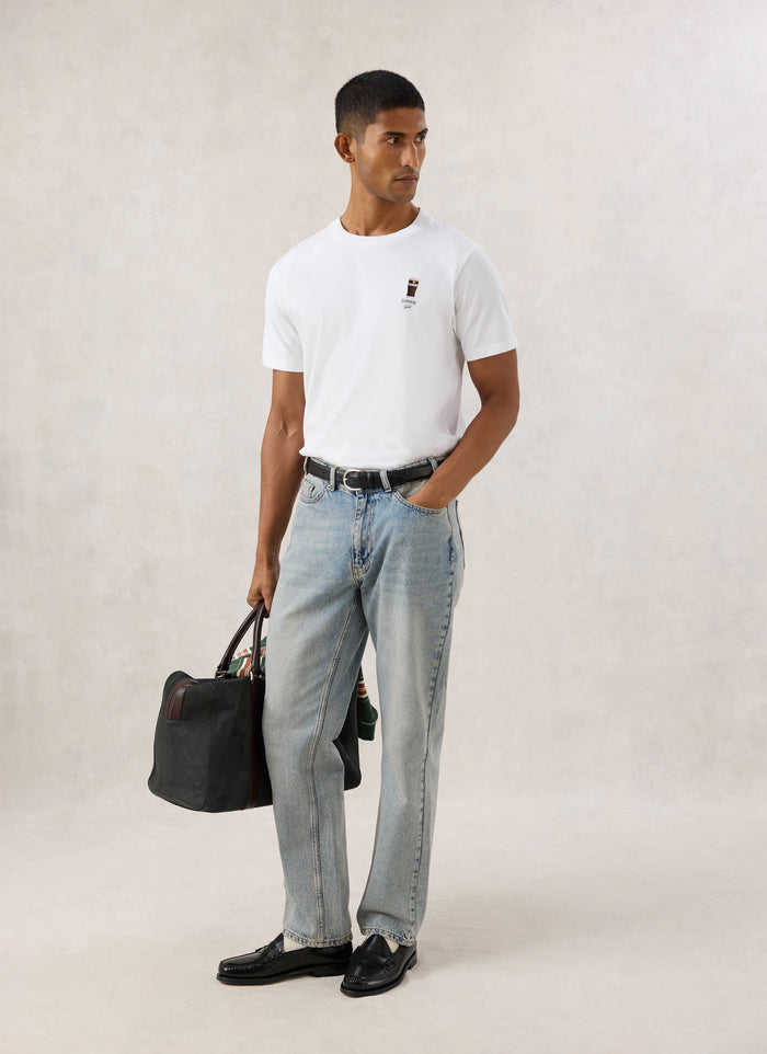 Model wearing the white Guinness x Percival stout embroidered T-shirt with light wash jeans and carrying a dark bag.