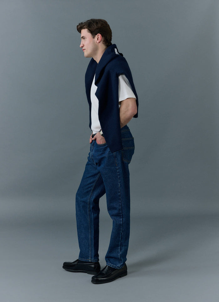 Model wears Percival mid-wash blue straight leg jeans with a white t-shirt, navy knit over his shoulders and black loafers.