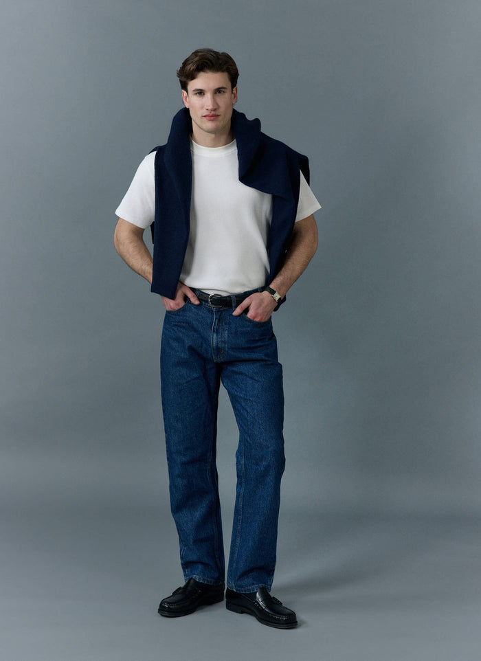 A man stands wearing Percival's mid wash blue straight leg jeans, a white t-shirt and a navy jumper over his shoulders.