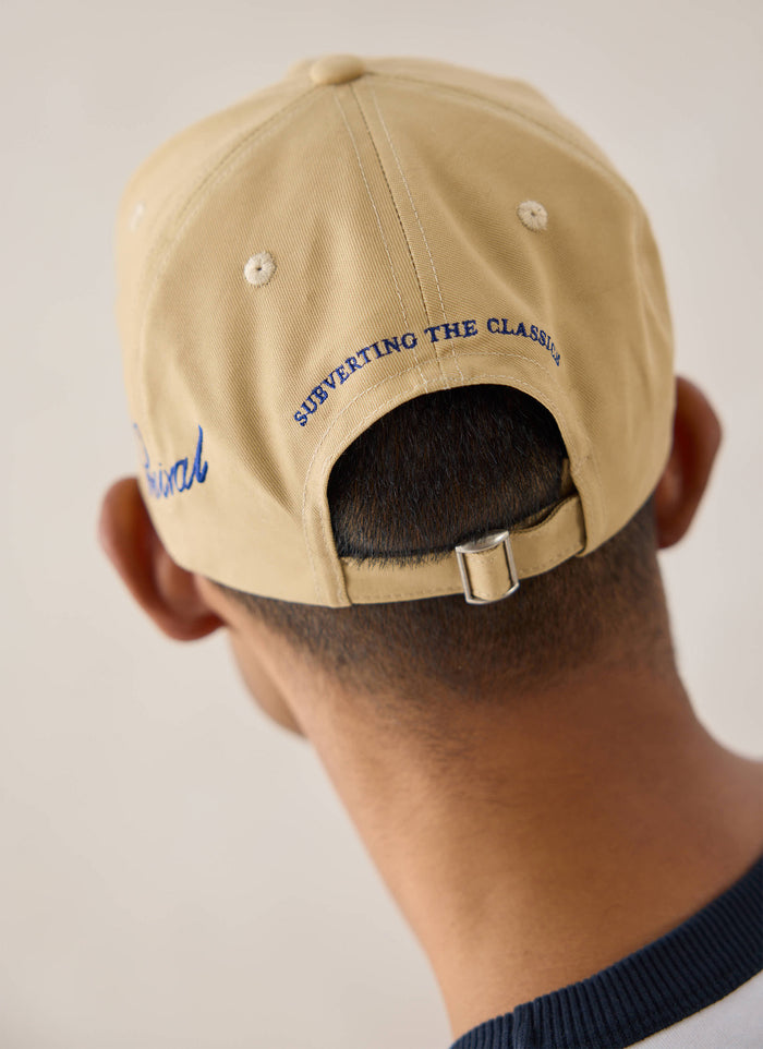 A close-up view of the back of the tan Percival trucker cap showing blue embroidery and strap closure.