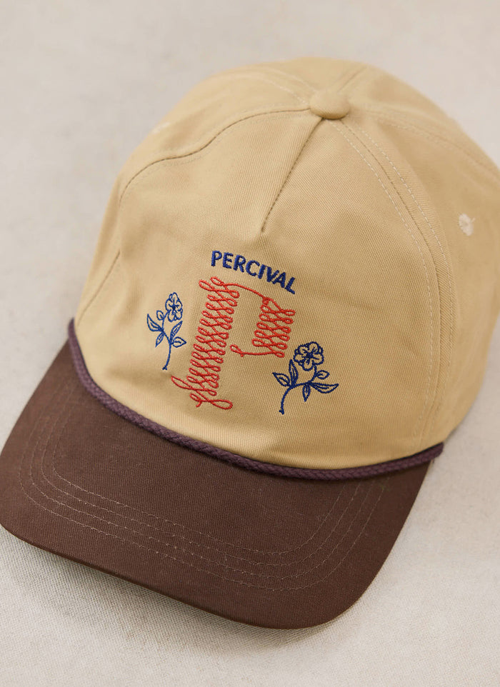 A close-up of the tan and brown Percival trucker cap, showing the embroidered floral and script details on the front.